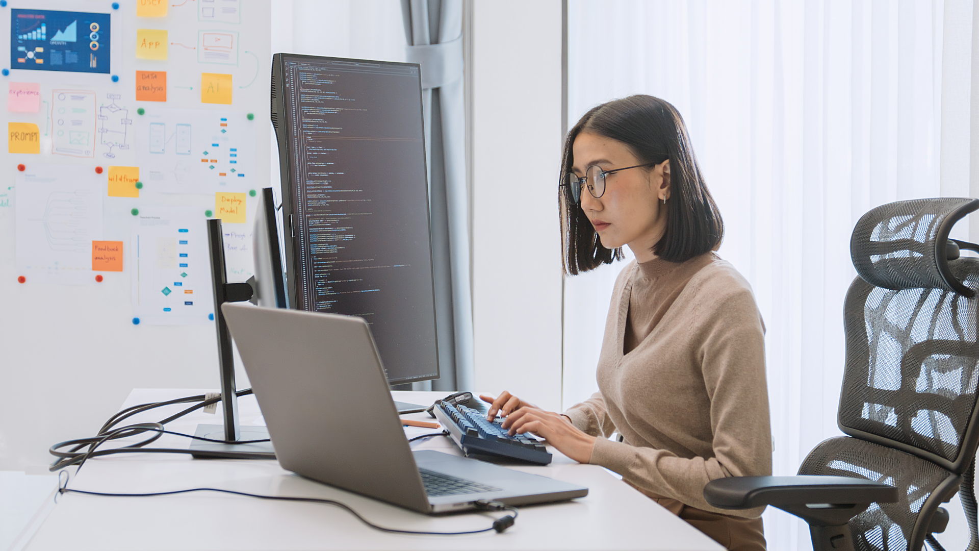 Female developer analyzing code on dual monitors beside sticky-note workflow diagrams