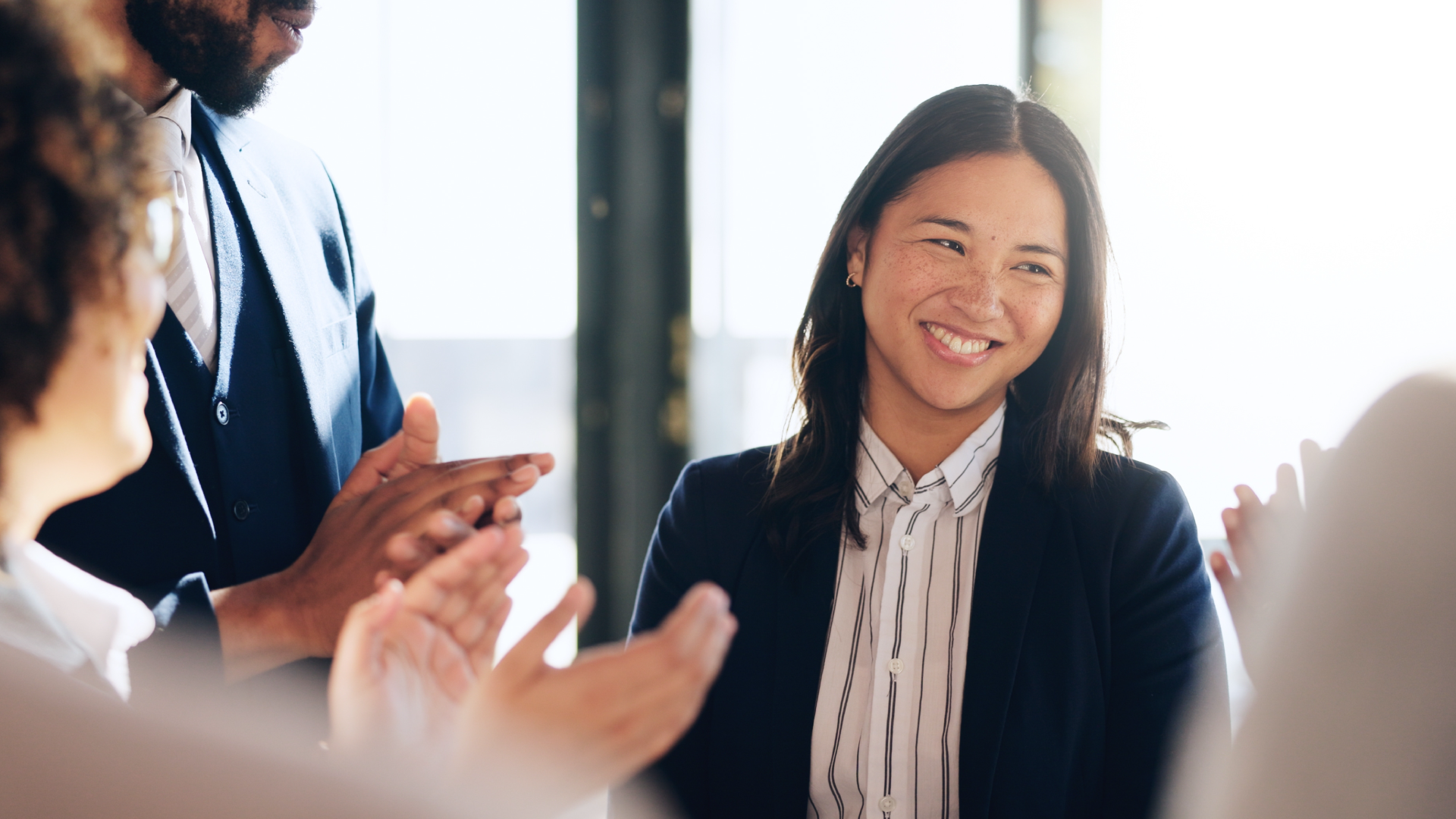 Smiling businesswoman in a navy blazer receives applause from colleagues in a bright office