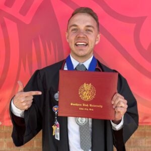 SUU online RN to BSN graduate Jackson McBride at graduation smiling and pointing to his diploma